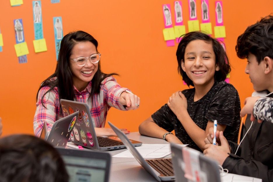 A teacher smiling and helping two students on their computer.